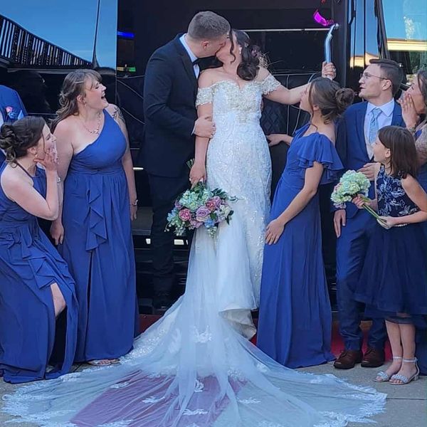 Bride and groom kiss surrounded by joyous wedding party in blue attire beside a black van.