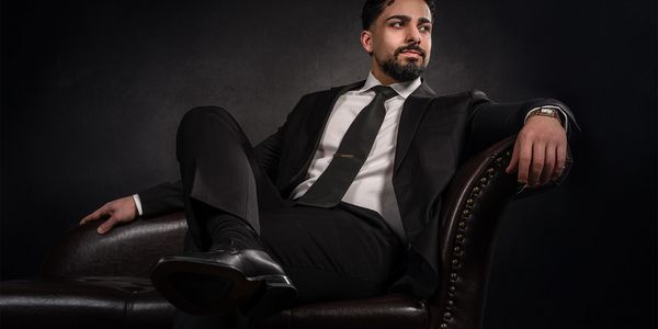 A confident man in a black suit lounging on a leather sofa against a dark background.