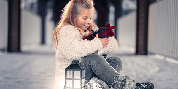 A young girl in winter clothes hugs a stuffed animal beside a lantern in the snow.