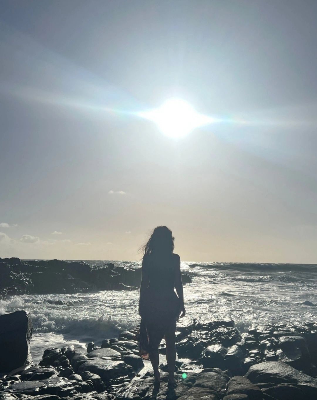 Sherridan looking out at the ocean, standing on deep black rocks, with a shining sun overhead