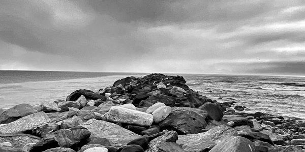 Black and white photo of rocky jetty extending into the ocean under cloudy sky.