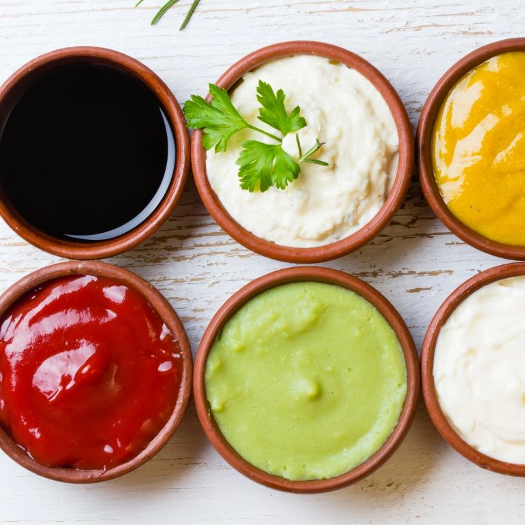 Six small bowls with different colorful sauces on a white wooden surface.