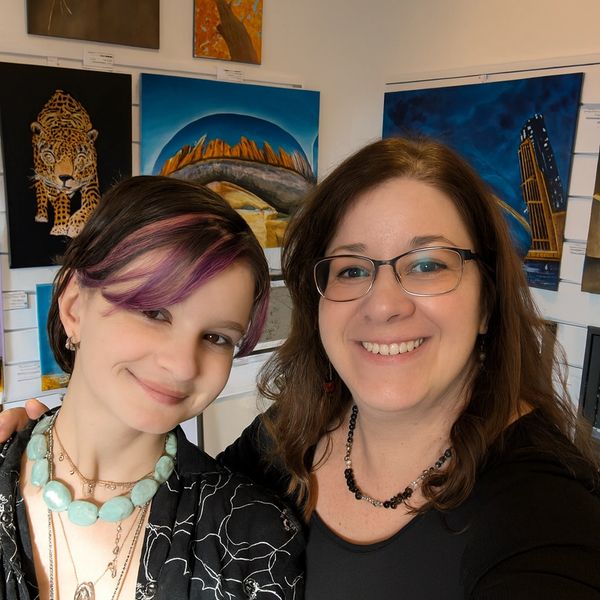 Two smiling women posing in an art gallery with colorful paintings behind them.
