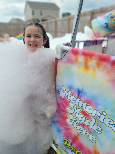 A smiling girl covered in bubbles next to a colorful sign.