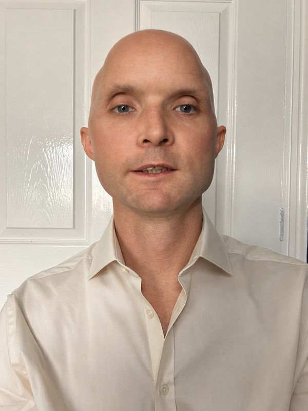 A bald man in a cream-colored shirt stands against a white door background.