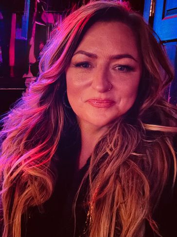 A woman with long wavy hair illuminated by pink and purple lighting.