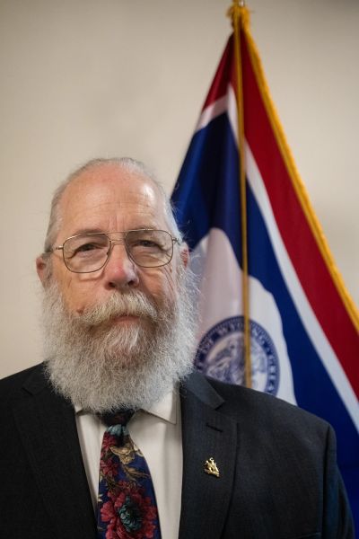 Bearded man in a suit with a colorful tie stands before a flag.