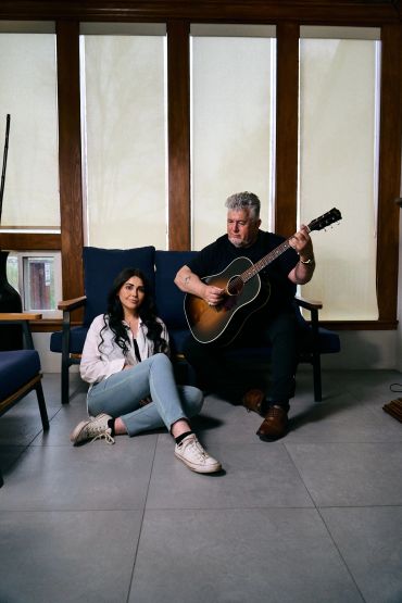 A man plays guitar while a woman sits on the floor nearby in a cozy room.