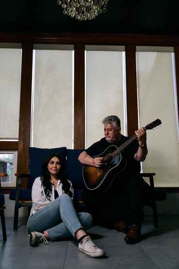 A man plays guitar while a woman sits on the floor beside him indoors.