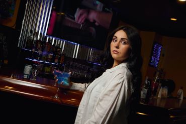 A woman in a white blouse holding a blue cocktail at a bar.