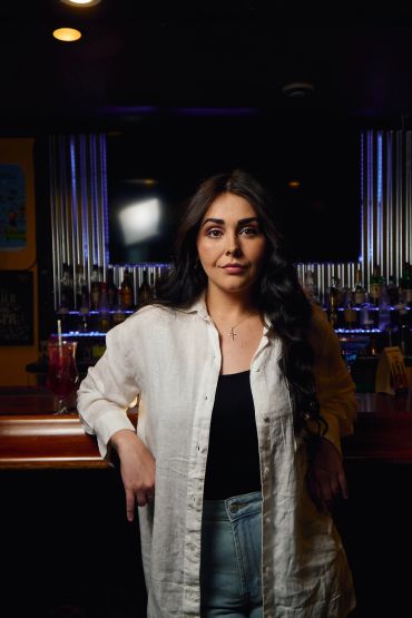 Confident woman standing in a dimly lit bar with arms resting on the counter.
