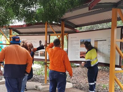 Workers in safety gear reading outdoor informational boards under shelter.