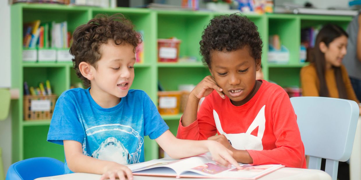 Two boys reading a book together at a table in a colorful classroom.