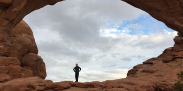 Person standing framed by a large rock arch under a cloudy sky.