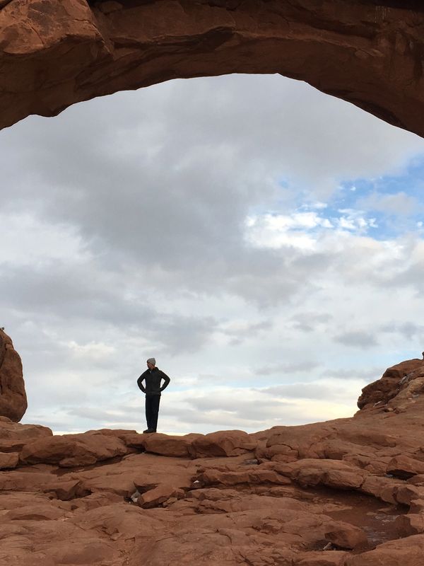 Person standing framed by a large rock arch under a cloudy sky.