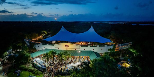 A lively outdoor event at dusk under large white tents surrounded by trees.