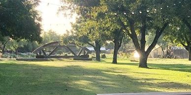 Sunlit park with trees and a small bridge in the background.