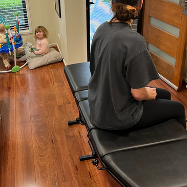 Young mum sitting on mobile chiropractic table with her young children sitting in the corner playing