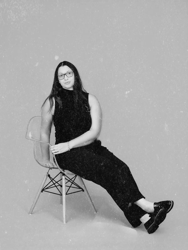 Woman in black outfit sitting on a transparent chair against a plain backdrop.