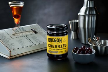 A jar of Oregon Fruit Co. cherries on a bar countertop with cocktail tools.