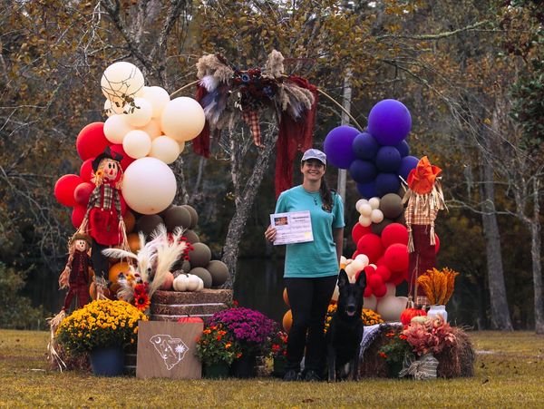 Woman with a dog holding a certificate in a colorful autumn-themed balloon arch setting.