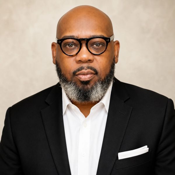 Portrait of a serious bald man with glasses and a beard in a black suit.
