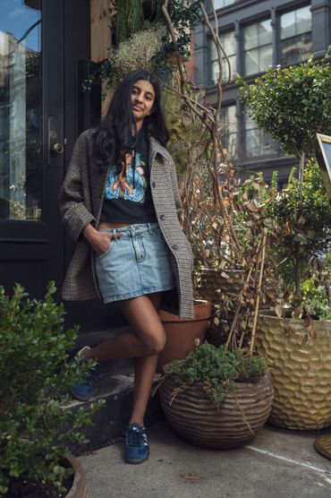 A young woman leans casually against a doorway surrounded by plants.