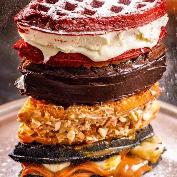 Stack of assorted waffles with various toppings and powdered sugar.