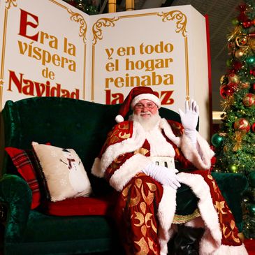Santa Claus waves while sitting on a green sofa beside a Christmas tree.