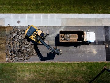 Aerial view of excavator loading debris into a dump truck on a paved road.