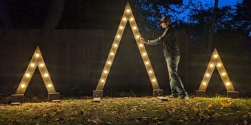 The CEO of The Starlight Project leaning against a hand-crafted, lighted, triangle wedding arch set.