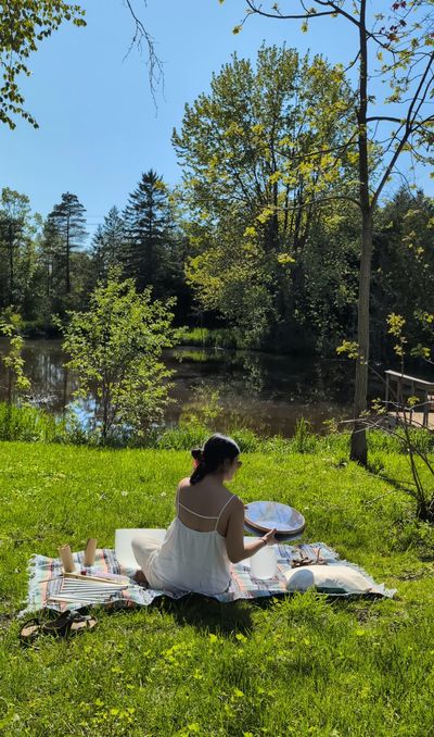 A woman in a white dress sits on a blanket near a peaceful pond surrounded by trees.