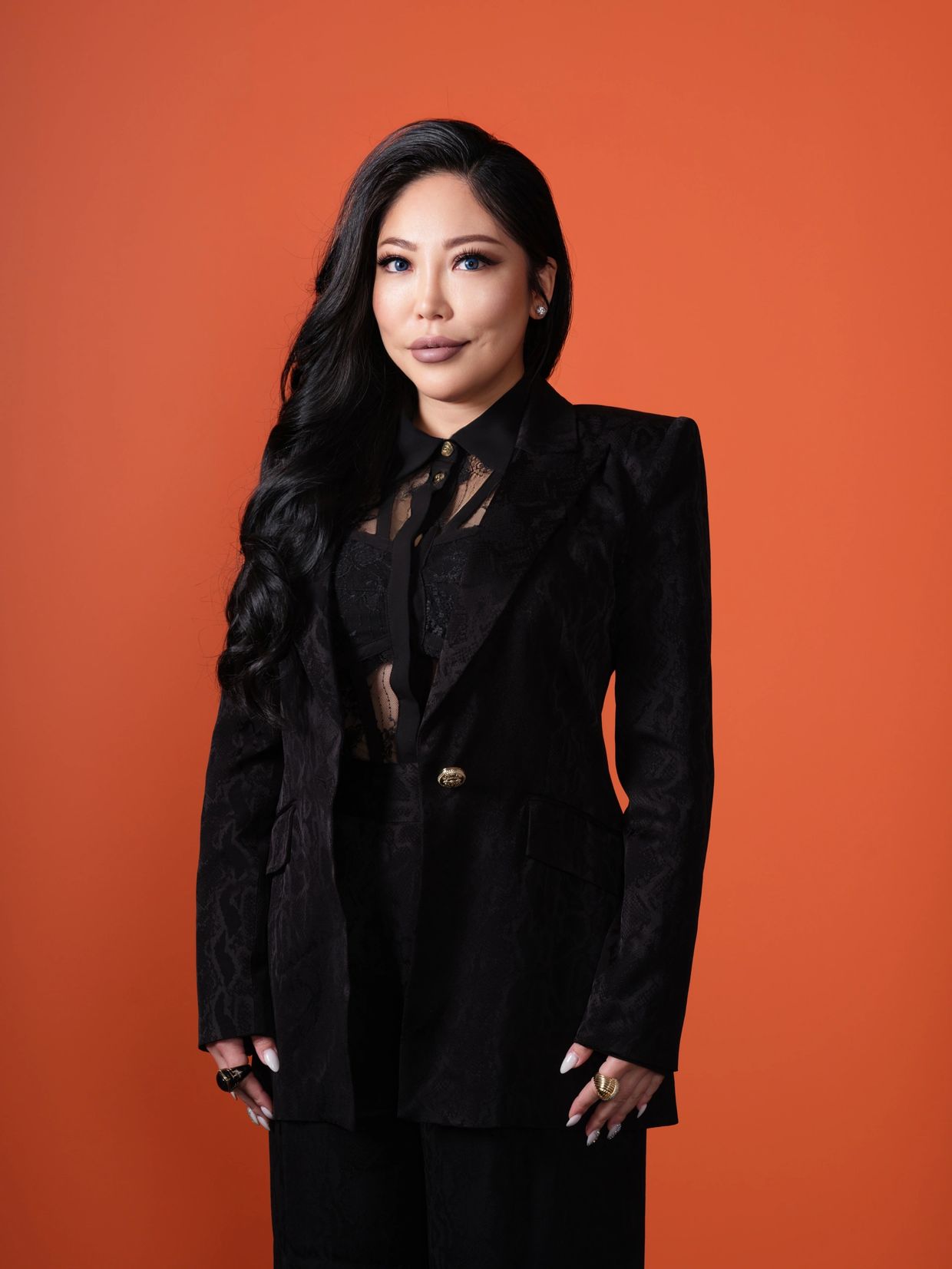 Confident woman in elegant black suit with long dark hair against an orange backdrop.