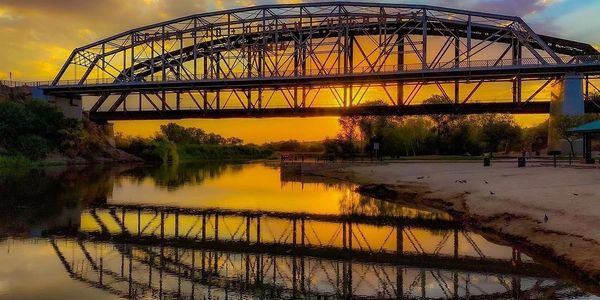 Steel bridge reflecting beautifully on calm river at sunset.