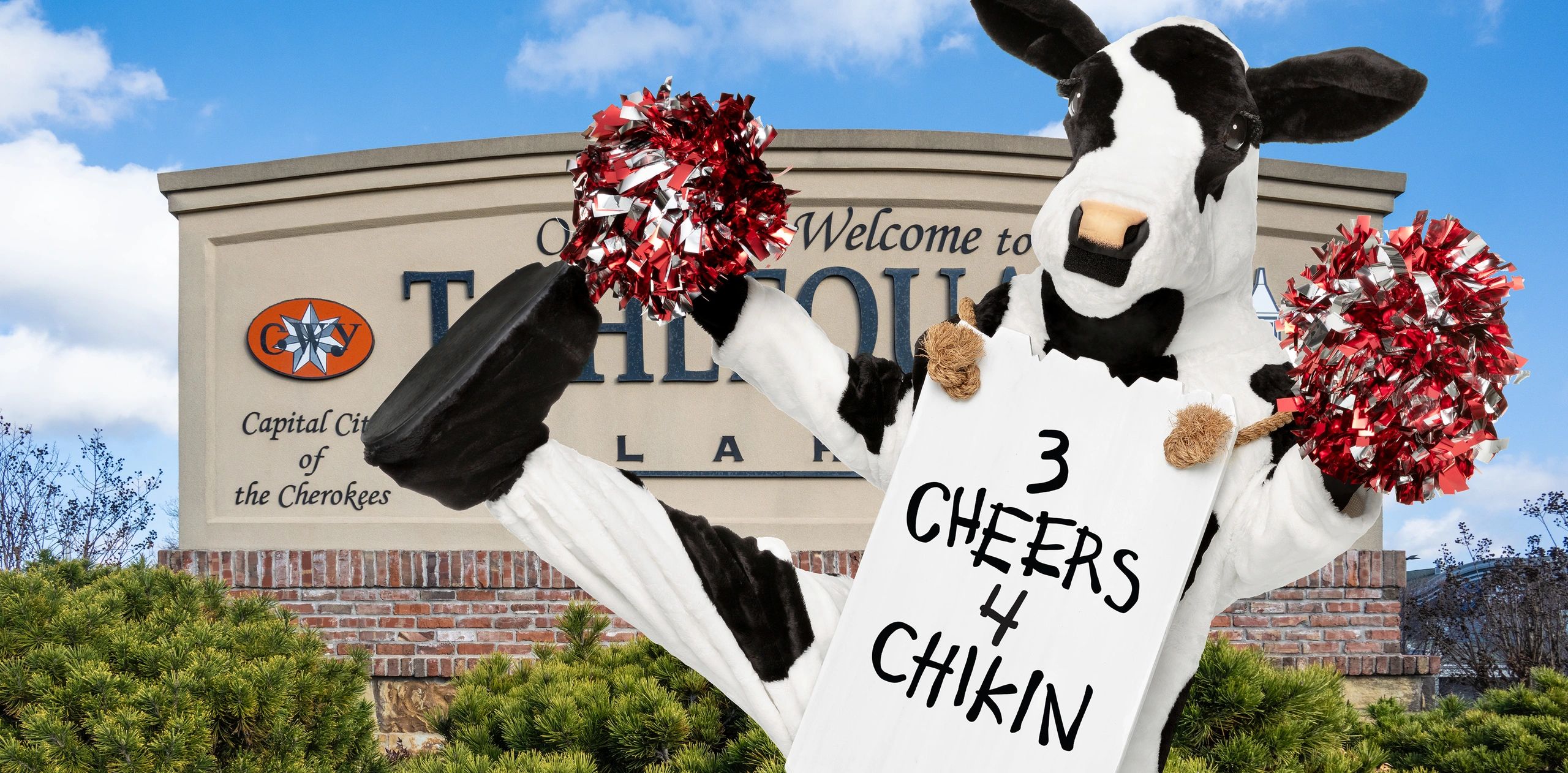 Cow mascot with pom-poms in front of a welcome sign.