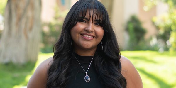 A smiling woman with long black hair and a pendant necklace outdoors.
