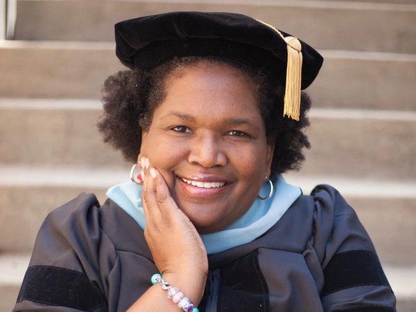Smiling graduate in cap and gown posing with hand on cheek.