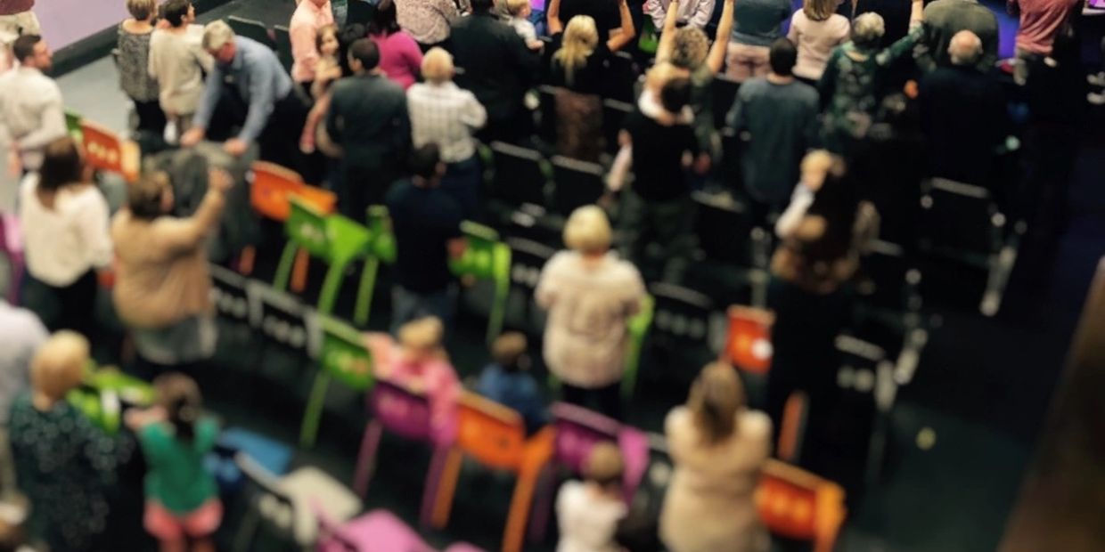 A crowded auditorium with people standing and colorful chairs.
