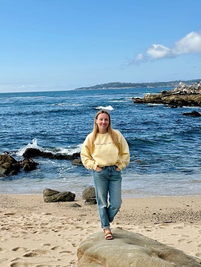 A woman stands on a rock at the beach with the ocean behind her.