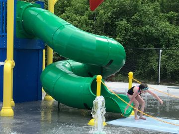 A child exits a green spiral water slide at a splash pad.