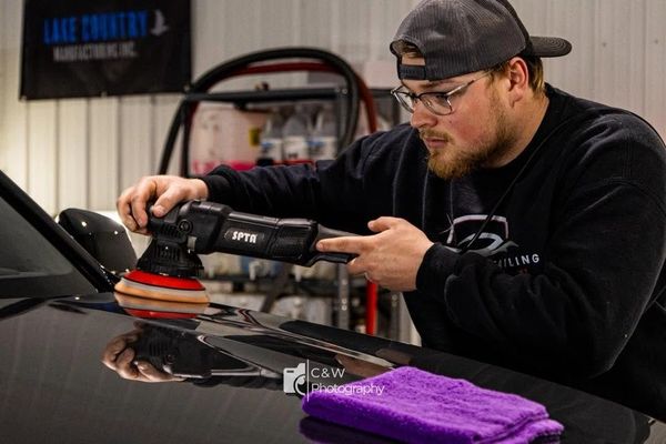 Man polishing a black car with a power buffer in a garage.