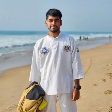 Young man in a white martial arts uniform stands on a beach holding a bag.