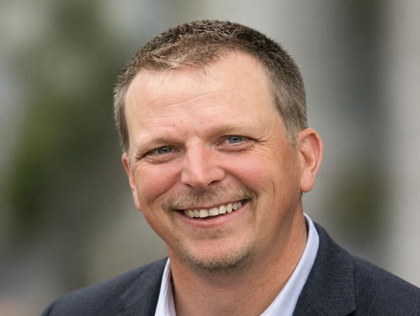 Smiling man in a suit jacket with a blurred outdoor background.