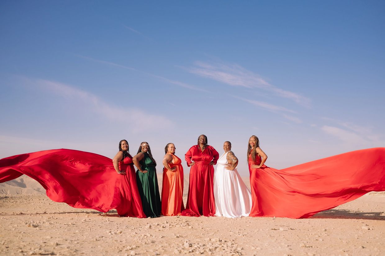 Six women in colorful flowing gowns pose confidently in a desert under a clear blue sky.