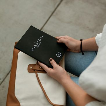 Person holding a black journal titled 'The Mission Journal' while sitting with a beige and brown bag.