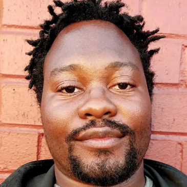 Close-up of a man with short dreadlocks and a goatee against a red brick wall.