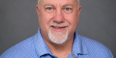 Smiling middle-aged man with a gray goatee wearing a blue patterned shirt.
