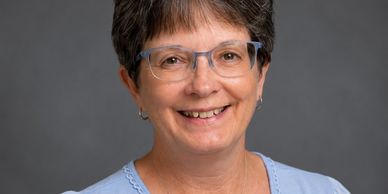Middle-aged woman with short hair and glasses smiling against a gray background.