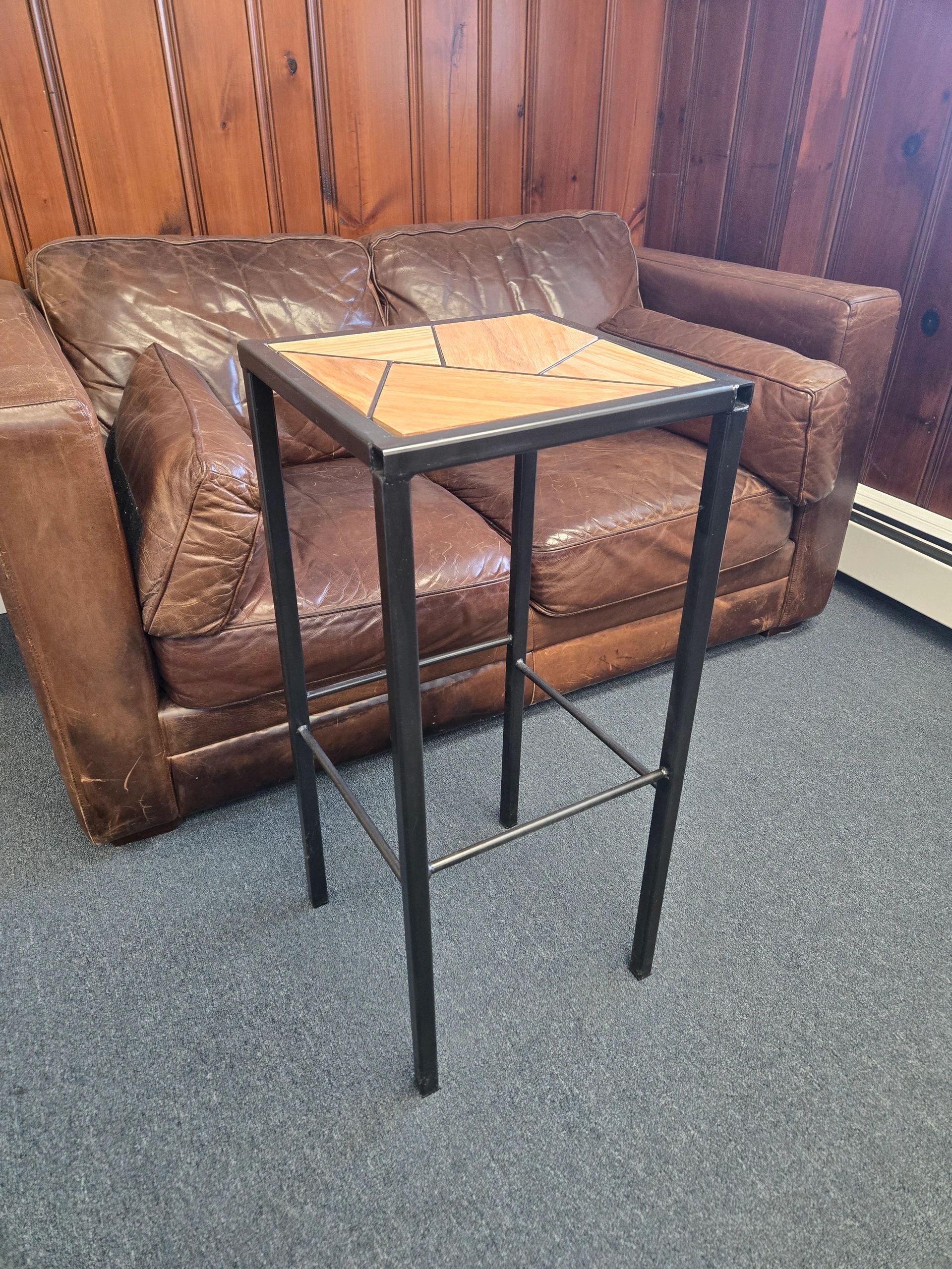 Tall metal table with wooden top in front of a brown leather couch.