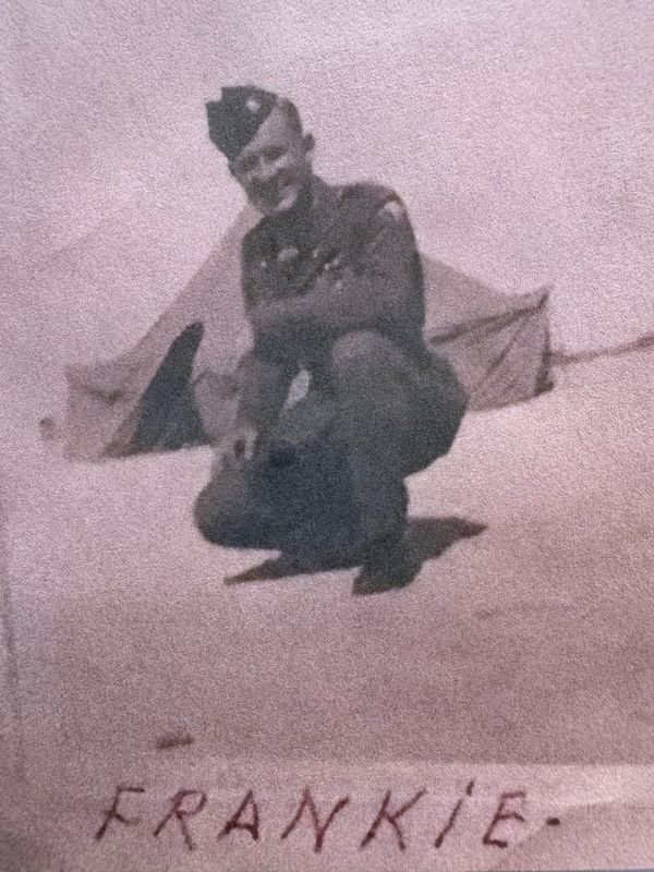 A soldier named Frankie squatting in front of a tent, smiling.
